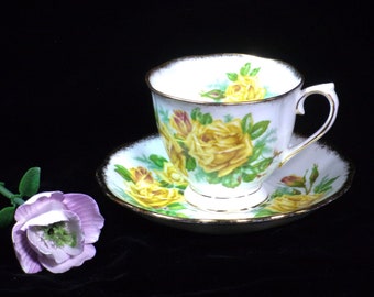 May include: A white teacup and saucer set with a gold rim and a yellow rose floral design. The teacup has a curved handle and sits on a matching saucer. A purple flower is in the foreground.