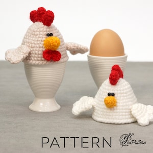 May include: Two crocheted chicken egg cosies, one in a white egg cup and one on a grey surface. The cozies are white with red combs, yellow beaks, and black eyes. The cozie in the egg cup has a red bow.