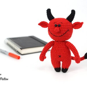 May include: A red crocheted devil plush toy with black horns and a tail. The toy has a smiling face with black eyes and a stitched-on mouth.