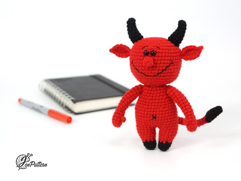 May include: A red crocheted devil plush toy with black horns and a tail. The toy has a smiling face with black eyes and a stitched-on mouth.