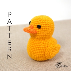 May include: A crocheted yellow rubber ducky toy with black eyes and an orange beak. The ducky is sitting on a beige surface.