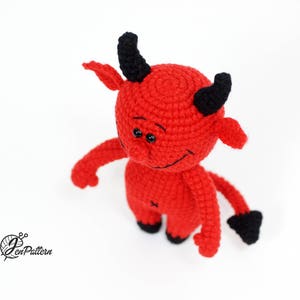 May include: A red crocheted devil plush toy with black horns and a tail. The toy has a friendly expression with big eyes and a small smile.
