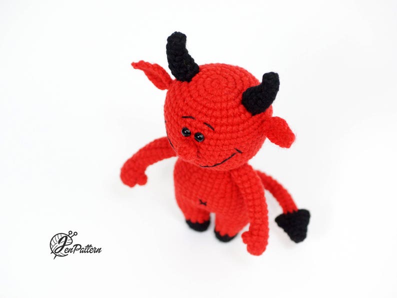 May include: A red crocheted devil plush toy with black horns and a tail. The toy has a friendly expression with big eyes and a small smile.