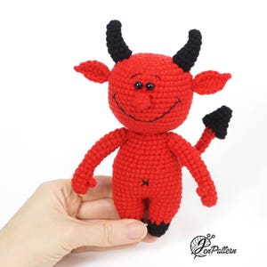May include: A red crocheted devil toy with black horns and a tail. The toy has a smiling face with black eyes and a stitched-on mouth.