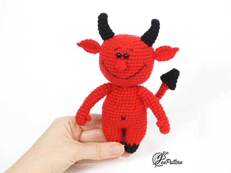 May include: A red crocheted devil toy with black horns and a tail. The toy has a smiling face with black eyes and a stitched-on mouth.