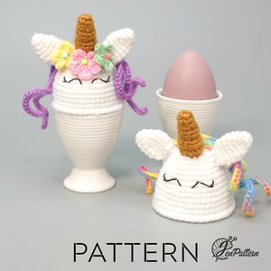 May include: Two crocheted unicorn egg cozies, one with purple yarn mane and flowers, the other with rainbow yarn mane. Both are white with brown horns and black eyes. The cozies are on white egg cups.