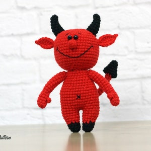 May include: A red crocheted devil plush toy with black horns, a tail, and a smiling face.