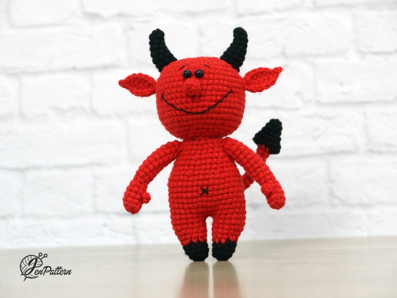 May include: A red crocheted devil plush toy with black horns, a tail, and a smiling face.