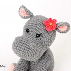 May include: A grey crocheted hippopotamus toy with pink ears and a pink flower with a yellow centre on its head. The hippopotamus has black eyes.