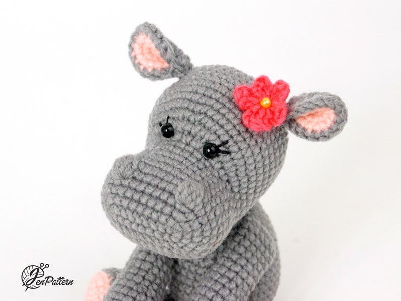 May include: A grey crocheted hippopotamus toy with pink ears and a pink flower with a yellow centre on its head. The hippopotamus has black eyes.