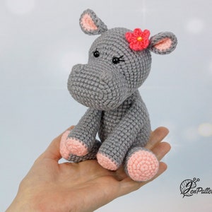 May include: A grey crocheted hippopotamus with pink ears and feet. The hippopotamus has a pink flower on its head.