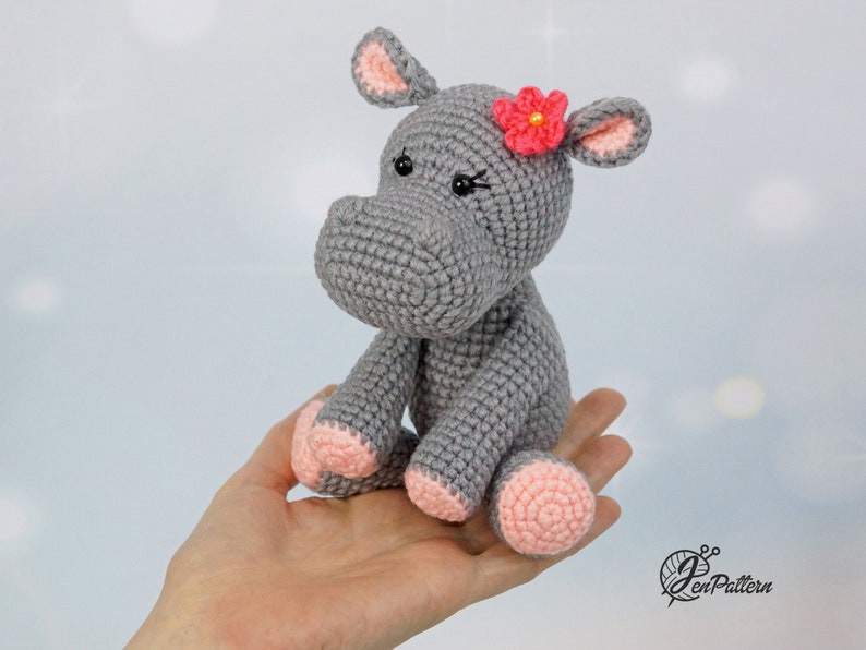 May include: A grey crocheted hippopotamus with pink ears and feet. The hippopotamus has a pink flower on its head.