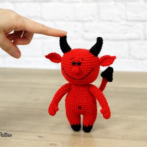 May include: A red crocheted devil toy with black horns and a tail. The toy has a smiling face with black eyes and a stitched-on mouth.