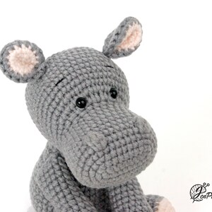 May include: Close-up of a grey crocheted hippopotamus toy with pink ears and black eyes. The toy is sitting on a white background.