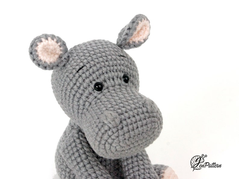 May include: Close-up of a grey crocheted hippopotamus toy with pink ears and black eyes. The toy is sitting on a white background.