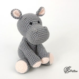 May include: A grey crocheted hippopotamus stuffed animal with pink ears and feet. The hippopotamus is sitting with its legs crossed.