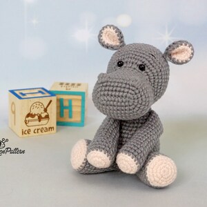 May include: A grey crocheted hippopotamus toy sitting on a wooden block with the words "ice cream" printed on it. The hippopotamus has pink ears and feet.