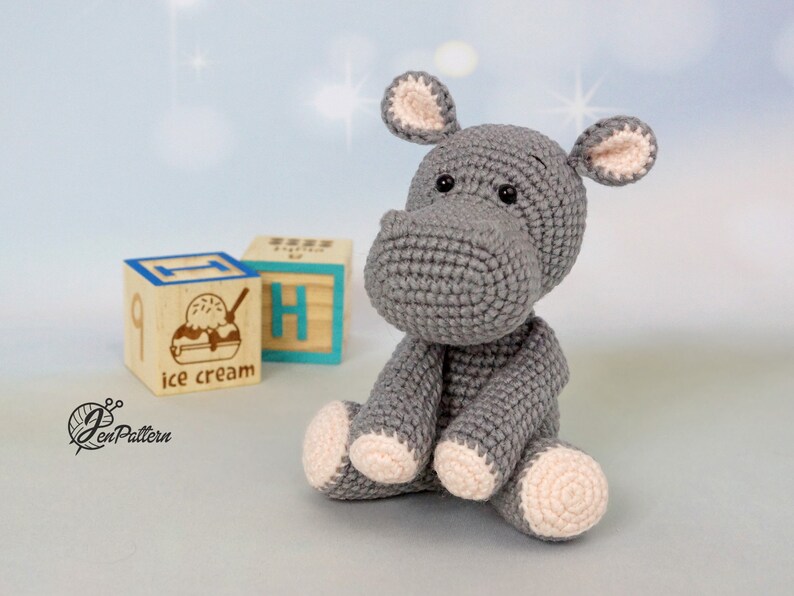 May include: A grey crocheted hippopotamus toy sitting on a wooden block with the words "ice cream" printed on it. The hippopotamus has pink ears and feet.