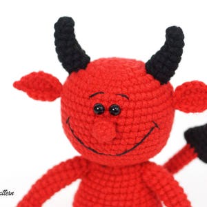 May include: A red crocheted devil with black horns and a tail. The devil has a smiling face with black eyes and a black stitched mouth.
