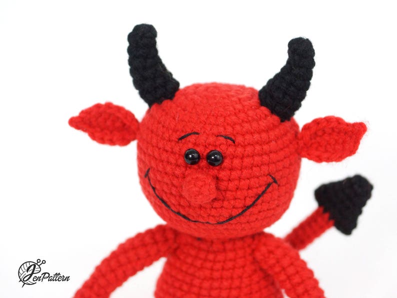 May include: A red crocheted devil with black horns and a tail. The devil has a smiling face with black eyes and a black stitched mouth.