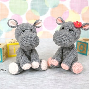 May include: Two grey crocheted hippopotamus toys sitting on a grey surface. One hippo has a pink flower on its head. Both hippos have pink feet.
