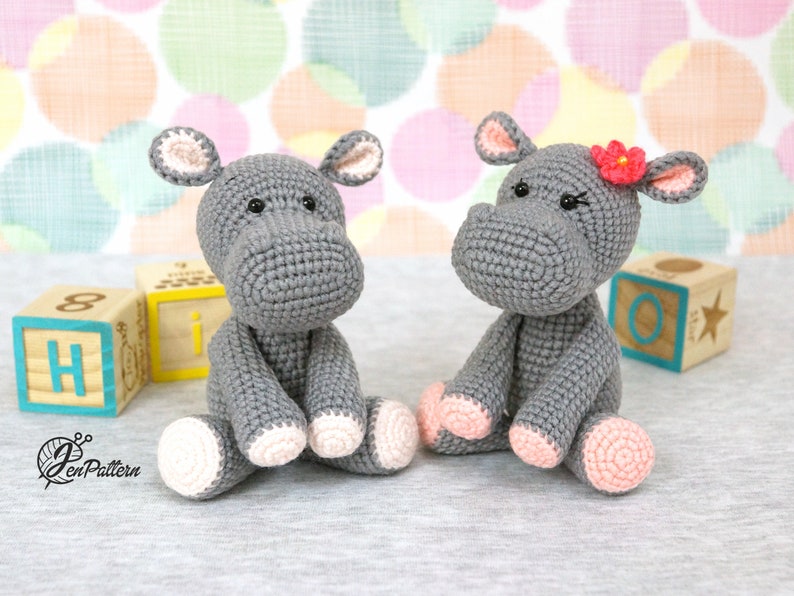 May include: Two grey crocheted hippopotamus toys sitting on a grey surface. One hippo has a pink flower on its head. Both hippos have pink feet.