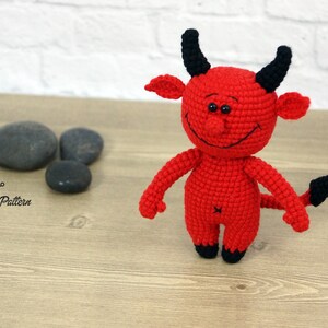 May include: A red crocheted devil toy with black horns, a tail, and eyes. The toy is smiling and has a stitched-on mouth.