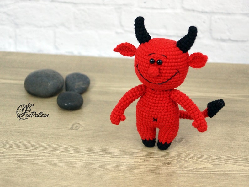May include: A red crocheted devil toy with black horns, a tail, and eyes. The toy is smiling and has a stitched-on mouth.