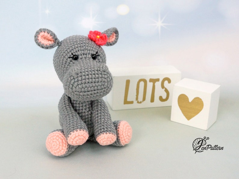 May include: A grey crocheted hippopotamus with pink feet and a red flower on its head. The hippopotamus is sitting on a white surface with two wooden blocks that say "LOTS" and a heart.