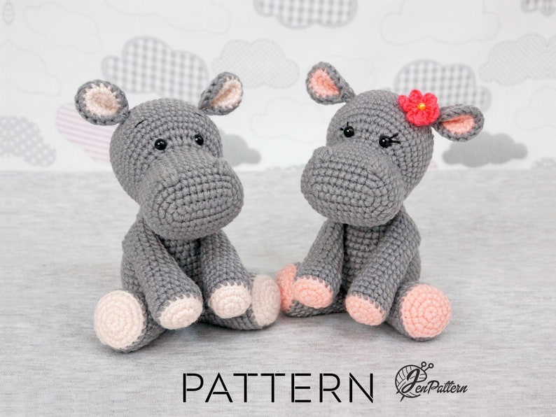May include: Two grey crocheted hippopotamus toys sitting on a grey surface. One hippo has a pink flower on its head.