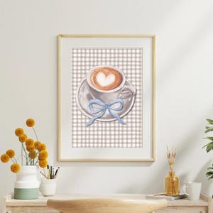 Könnte beinhalten: Gerahmter Kunstdruck einer Kaffeetasse mit herzförmigem Design im Schaum, auf einer Untertasse mit hellblauer Schleife. Der Hintergrund zeigt ein kariertes Muster in Braun und Weiß. Der Rahmen ist aus hellem Holz.