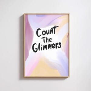 以下が含まれることがあります： 「Count The Glimmers」の文字が黒い筆記体で書かれた、額装されたアートプリント。背景は、ピンク、紫、黄色の柔らかな水彩画。フレームは明るい木の色。