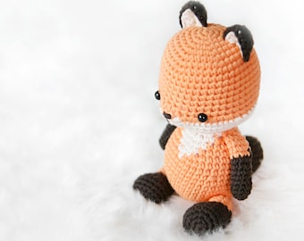 May include: A crocheted fox plush toy with orange fur, black ears, and black eyes. The toy is sitting on a white fluffy surface.