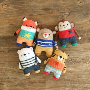 May include: Five crocheted stuffed animals in various colors and designs. The animals include a fox, a reindeer, a monkey, a polar bear, and a tiger. The animals are all sitting on a wooden surface.