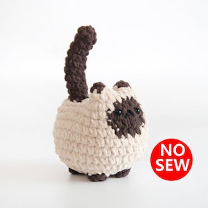 May include: A crocheted stuffed cat toy with a brown tail and a cream-colored body. The cat has a brown face and black eyes. The toy is labeled "NO SEW".