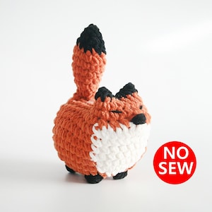 Op de afbeelding: Een gehaakt vosje met een witte buik en een zwarte staartpunt. Het speelgoed is gemaakt van oranje, wit en zwart garen. Het speelgoed ligt op een wit oppervlak. De tekst "NO SEW" staat in rood op een witte cirkel.
