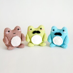 May include: Three crocheted frog plushies with white bellies and black mustaches. The frogs are brown, green, and blue.