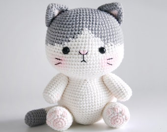 May include: A crocheted gray and white cat plush toy with pink accents on the paws and nose. The cat is sitting with its tail curled behind it.