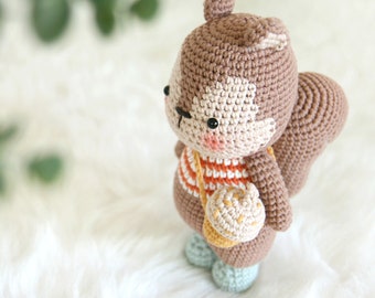 Könnte beinhalten: Ein braunes Häkel-Eichhörnchen mit einem weiß-orange gestreiften Shirt und einer braunen Häkel-Eichel in der Hand. Das Eichhörnchen steht auf einer weißen Oberfläche.