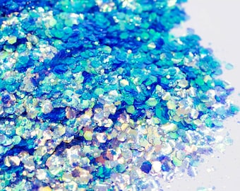 May include: A close-up of iridescent blue and silver glitter. The glitter is scattered on a white surface.