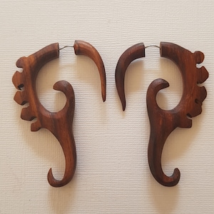 May include: A pair of wooden ear gauges with a unique, intricate design. The gauges are brown and have a natural wood finish. They are made of a single piece of wood and have a curved shape.