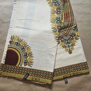 Könnte beinhalten: Cremefarbener Stoff mit aufwendigen Mustern in Gelb, Blau, Grün und Burgunderrot. Das Design zeigt geometrische Formen und florale Motive mit einem Rand aus sich wiederholenden pfeilartigen Mustern. Der Stoff trägt die Aufschrift "Supreme Wax Holland" und "SWA 0764".