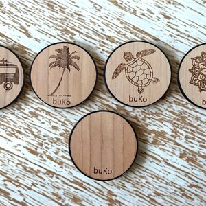 Puede incluir: Cinco posavasos redondos de madera con borde negro. Cada posavasos presenta un diseño diferente: una palmera, una tortuga, un mandala, una furgoneta vintage y un posavasos liso. Todos los posavasos están etiquetados con la palabra "buKo".