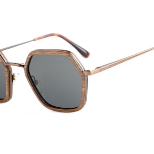 Puede incluir: Gafas de sol de forma octogonal con montura de madera y lentes grises oscuros. La montura es de color marrón oscuro con un acabado metálico.