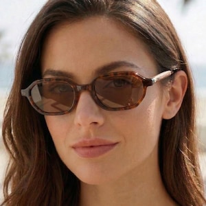 Puede incluir: Gafas de sol ovaladas con estampado de carey. Las gafas tienen lentes tintadas marrones y una montura marrón oscuro. La imagen es un primer plano.