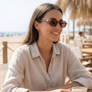 Puede incluir: Una persona con gafas de sol marrones con montura de carey y una camisa clara abotonada. Está sentada en una mesa al aire libre, con una playa y el mar de fondo. Las gafas de sol tienen forma redondeada.