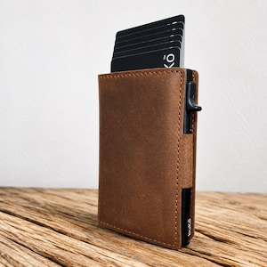 May include: A brown leather card holder wallet with a vertical black metal card ejection mechanism on the side. Several black credit cards are partially extended from the top, with the text "buKō" visible on the lower right side of the frame.