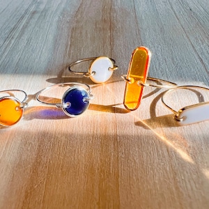 May include: Five gold rings with different coloured glass inlays. The rings feature a round orange, round blue, round white, rectangular orange, and rectangular white inlay.