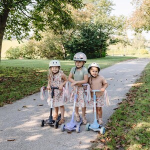 May include: Three children wearing helmets ride colourful scooters with tassels on a paved path. The scooters have three wheels and are decorated with floral patterns.