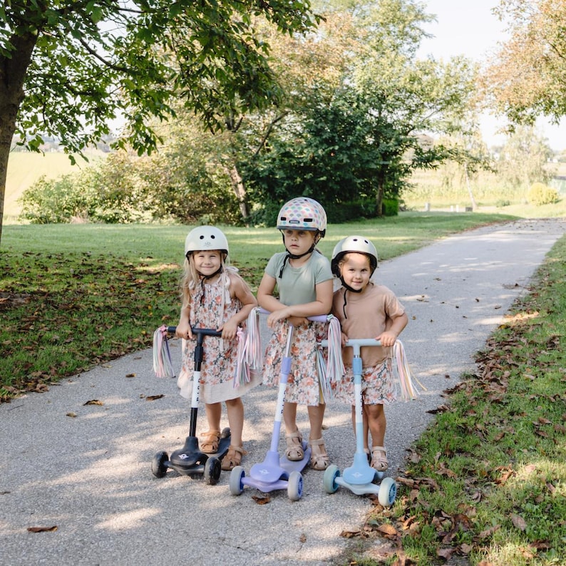 May include: Three children wearing helmets ride colourful scooters with tassels on a paved path. The scooters have three wheels and are decorated with floral patterns.